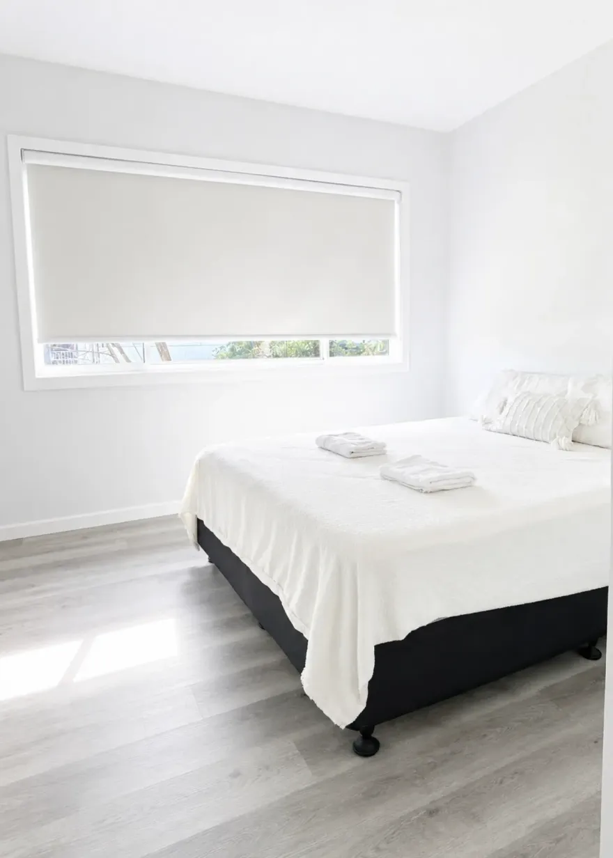 Bedroom with queen bed, white bedding and dark base beside a wide roller-blind window