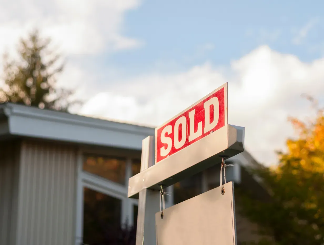 A red SOLD sign in front of a modern house under a blue sky with autumn trees